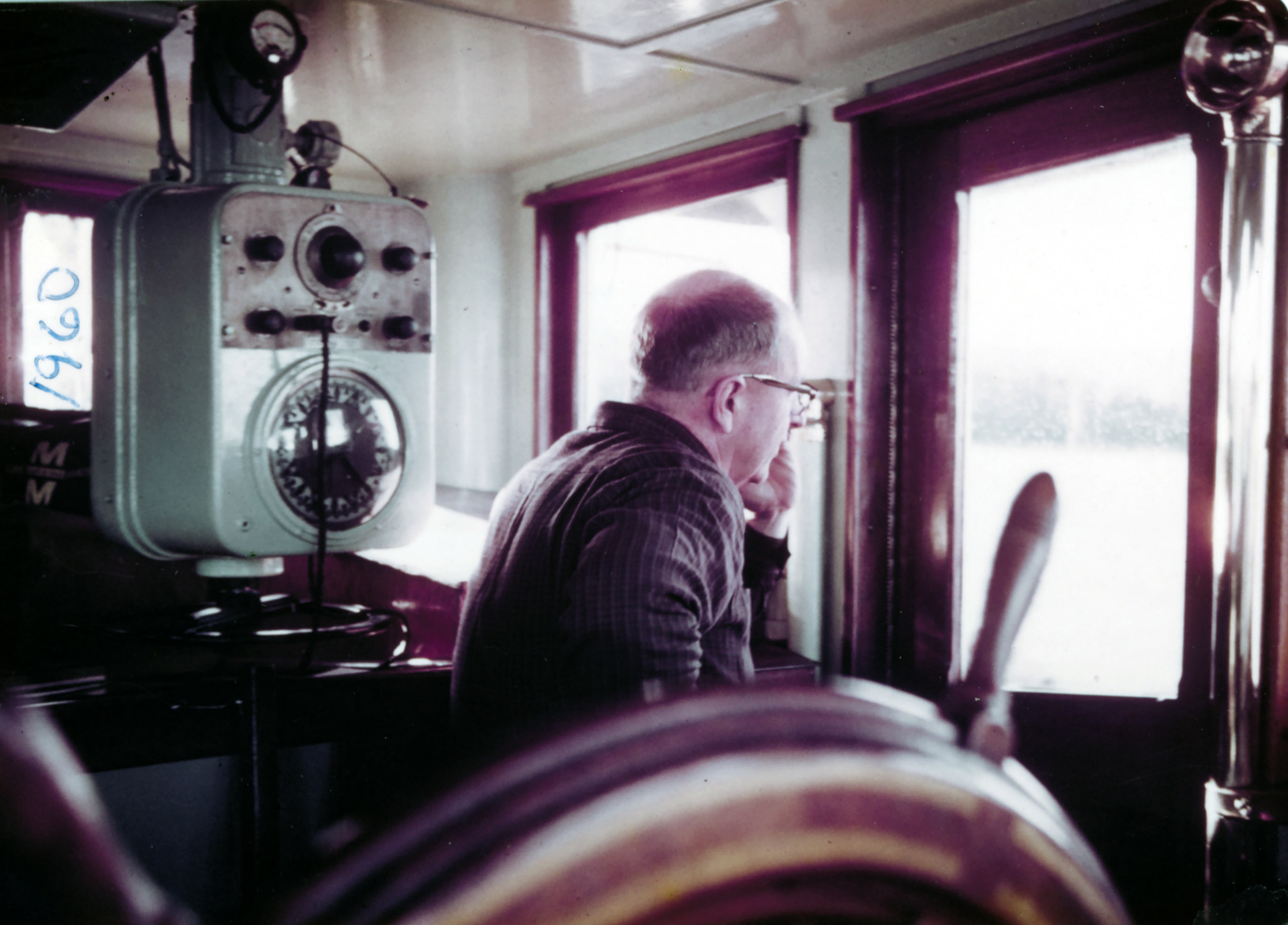 A man standing in a pilot house near a ship's wheel looking out the window A man standing in a pilot house near a ship's wheel looking out the window